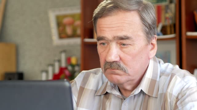 An-elderly-man-with-a-mustache-sits-behind-a-laptop-and-solves-problems.-He-looks-seriously-at-the-monitor