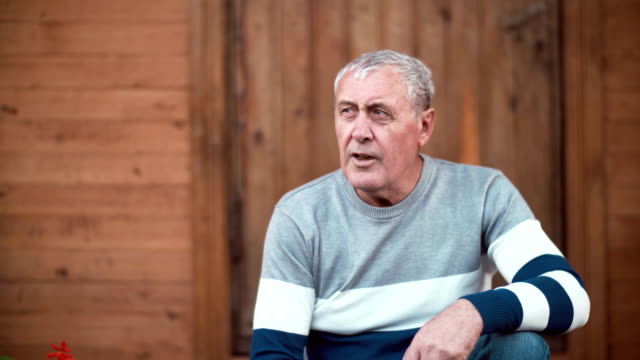 Old-man-with-gray-hair-sitting-in-front-of-camera-and-taking-conversation-with-somebody.-Wooden-background.-4K