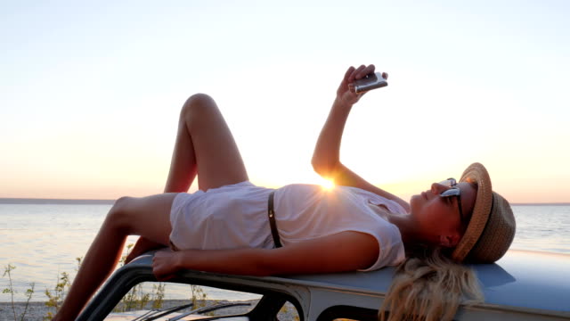 Happy-Journey,-young-woman-lying-on-rooftop-auto-makes-pictures-on-phone,-female-make-selfie-photo-at-mobile-on-retro-car