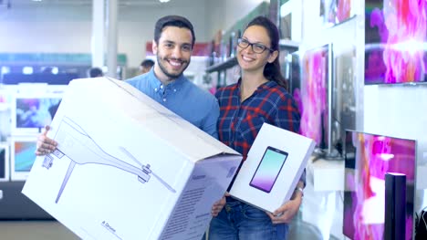 In-the-Electronics-Store-Happy-Young-Couple-Poses-with-Newly-Purchased-Drone-and-Tablet-Computer.-Store-is-Big-and-Bright,-Has-All-the-Latest-TV's,-Cameras-and-Smartphones.