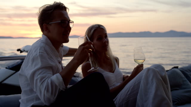 Young-Couple-Talk,-Drink-Champagne-in-the-Stern-of-the-Moving-Yacht.-They-Have-Great-Romantic-Evening.-In-the-Background-Island-with-Small-Village.