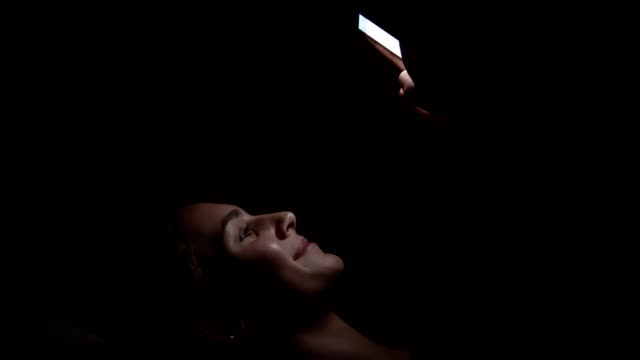 Mujer-sonriente-joven-tendido-en-un-sofá-y-usar-su-teléfono-en-una-habitación-oscura,-charlando-en-la-oscuridad-con-smartphone,-comunicarse-con-amigos-en-línea