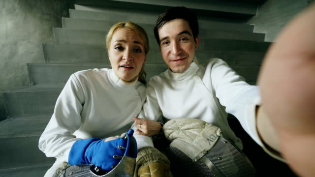 POV-of-Two-young-fencers-man-and-woman-have-online-video-call-with-trainer-using-smartphone-camera-after-fencing-competition-indoors