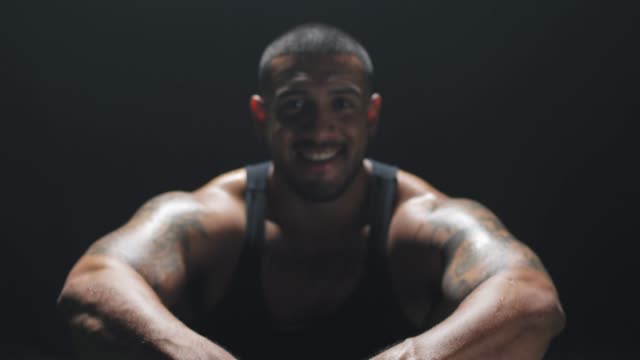 Zooming-shot-of-a-muscular-black-man-sitting-down-smiling-at-the-camera-on-a-dark-background