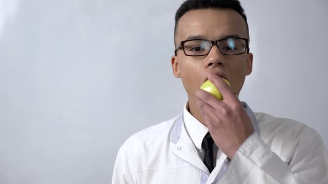 Young-successful-African-scientist-in-a-white-lab-coat-and-glasses-bites-an-apple,-eats-and-looks-at-the-camera,-portrait-concept.-60-fps