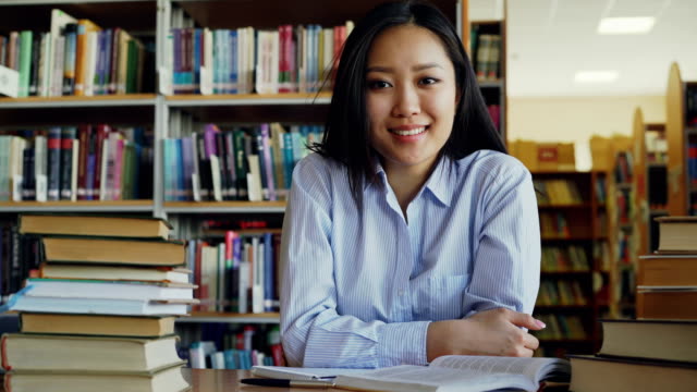 Porträt-des-jungen-schöne-asiatische-Studentin-mit-Haufen-von-Lehrbüchern-in-Bibliothek-Blick-in-die-Kamera-am-Tisch-sitzen.-Sie-lächelt-positiv.