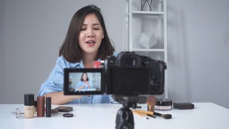 Glücklich-lächelnd-asiatische-Frau-oder-Beauty-Blogger-mit-Pinsel-und-Kamera-Videoaufnahme-und-winkenden-Hand-zu-Hause.-Videoblog-Blogging-Menschen-Schönheitskonzept.-Dolly-erschossen.