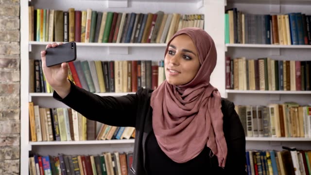 Young-muslim-beautiful-women-in-hijab-taking-selfie-then-looking-at-phone-and-watching-photos-in-library,-smiling