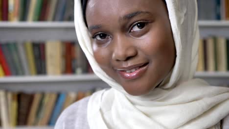 Face-of-young-charming-african-muslim-girl-in-hijab-is-watching-at-camera,-religioun-concept,-booksheves-on-background