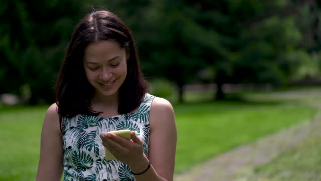Close-up-of-a-happy-looking-asian-woman-walking-in-the-park-and-typing-sms-on-the-yellow-smartphone.