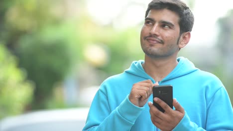 Young-happy-Persian-man-thinking-while-using-phone-outdoors
