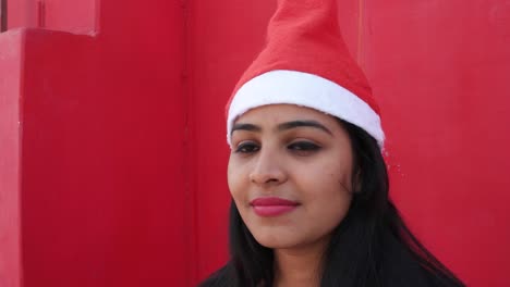 Young-Indian-woman-with-Santa's-hat-welcome-and-making-hand-gestures,-excited-and-super-happy,-with-a-matching-red-background,-handheld-gimbal-stabilized