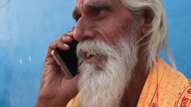 Tilt-up-to-Sadhu,-Hindu-priest,-talking-on-smart-phone-with-a-blue-background