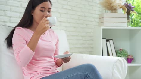 Mujer-asiática-joven-alegre-beber-café-caliente-o-té-disfrutando-mientras-está-sentado-en-su-sala-de-estar-en-casa.-Atractiva-mujer-asiática-feliz-sosteniendo-una-taza-de-café.