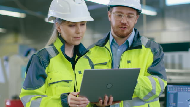 Male-and-Female-Industrial-Engineers-Wearing-safety,--Work-on-a-Manufacturing-Plant,-They-Discuss-Project-while-Using-Laptop.In-the-Background-Industrial-Facility.