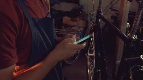 theme-small-business-bike-repair.-Young-caucasian-brunette-man-wearing-safety-goggles,-gloves-and-fartukhe-uses-mobile-phone-technology,-takes-notes,-checklist-in-bicycle-workshop