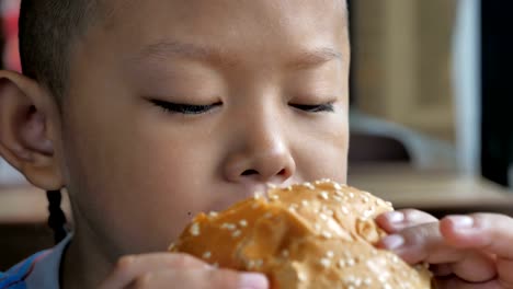 Cerca-niño-asiáticos-comer-hamburguesa,-lindo-muchacho-feliz-con-hamburguesa-en-el-restaurante.-Video-Slow-motion