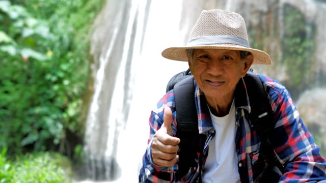 Senior-man-asian-hand-with-thumb-up-sit-in-front-of-the-waterfall.-Success,-Lifestyle-Travel-Adventures-and-tourism--with-backpack.-Video-Slow-motion