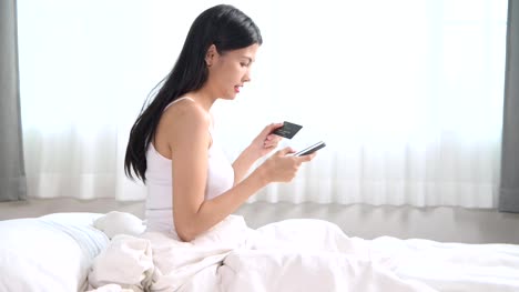 Mujer-de-compras-en-la-cama.-Mujer-asiática-mediante-teléfono-móvil-sentado-en-la-cama-a-la-tienda-en-línea-y-clave-de-tarjeta-de-crédito.-Concepto-de-compras-en-línea.