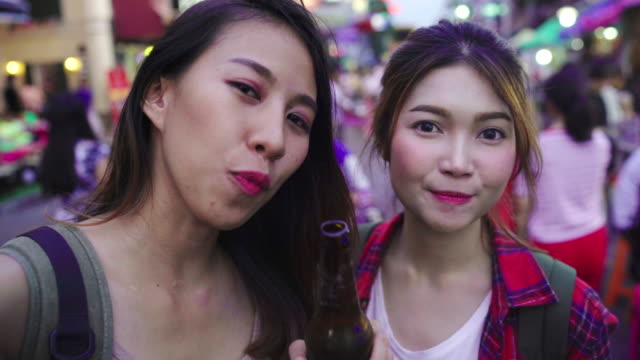 Viajero-mochilero-blogger-par-de-lesbianas-lgbt-de-Asia-las-mujeres-viajar-con-smartphone-para-selfie-en-Tailandia.-Mujer-bebiendo-alcohol-o-cerveza-en-Khao-San-Road,-la-calle-más-famosa-de-Bangkok.