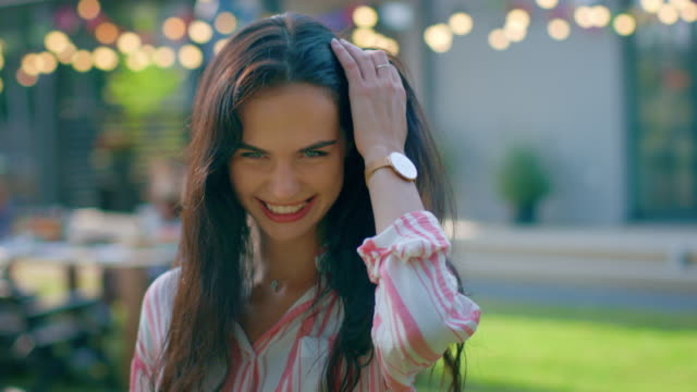 Portrait-of-Beautiful-Brunette-with-Loose-Hair-Charmingly-Smiling,-Straightening-Her-Hair.-In-the-Background-Hot-Summer-Day-and-Garden-Party-in-the-Backyard.-In-Slow-Motion.