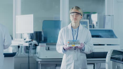 Weibliche-Wissenschaftler-führt-durch-Labor-mit-Tablett-mit-Reagenzgläsern-mit-Proben-gefüllt.-Im-Hintergrund-Mitarbeiter-im-Labor-mit-innovativen-Geräten.-In-Zeitlupe.