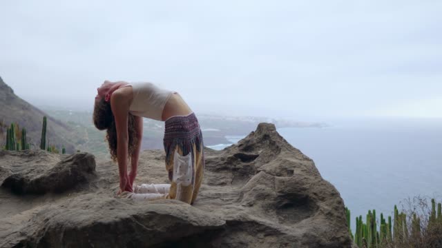 Chica-practicando-yoga-en-las-rocas-contra-el-cielo-azul-y-el-azul-del-mar.-Mujer-está-parado-en-una-piedra-en-una-postura-de-puente.