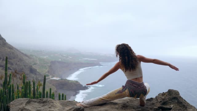 Eine-Frau-sitzt-auf-dem-Rand-einer-Klippe-in-eine-Pose-Krieg-mit-Blick-auf-den-Ozean-erheben-ihre-Hände-und-atmen-Sie-die-frische-Seeluft-dabei-yoga