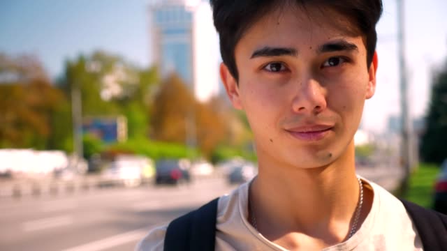 Close-up-beutiful-face-og-asian-guy-looking-at-camera-relaxed-and-standing-near-highway,-city-life,-warm-sunny-day