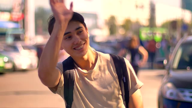 Happy-surprised-asian-boy-is-waving-on-the-street,-while-staying-near-road-with-cars-on-background