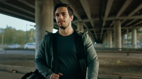 Portrait-shot-of-an-Athletic-Young-Man-Walking-From-the-Road-Under-a-Bridge-in-an-Urban-Environment.-He's-Wearing-a-Grey-Hoodie-and-a-Sports-Bag.