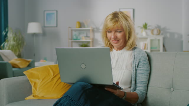 Portrait-of-a-Beautiful-Middle-Age-Woman-Relaxes-on-a-Sofa-in-Her-Living-Room,-Uses-Laptop.-Woman-Browses-Through-Internet-and-Doing-Work-from-Home-Office.