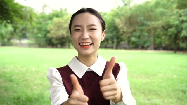 Joven-atractiva-mujer-asiática-en-uniforme-mostrando-los-pulgares-para-arriba-gesto-en-el-parque-al-aire-libre,-como-gesticular-de-la-escuela