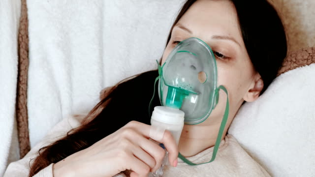Utilice-el-nebulizador-y-el-inhalador-para-el-tratamiento.-Mujer-joven-inhalando-a-través-de-mascarilla-inhalador-tumbado-en-el-sofá.-Vista-lateral.