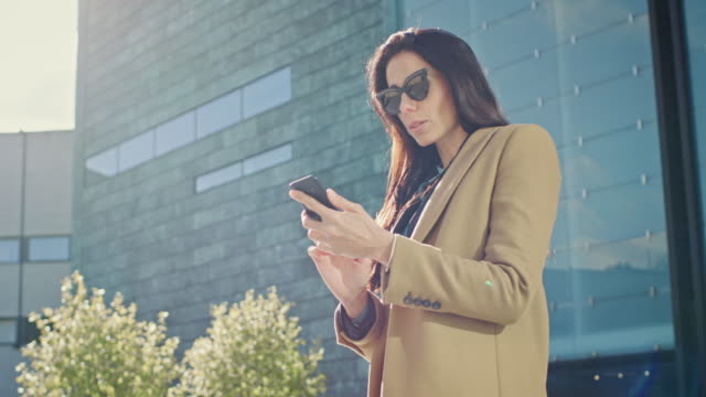 Elegant-Businesswoman-Uses-Smartphone-to-Conduct-Business-while-Standing-Near-Modern-Glass-Building.-Beautiful-Stylish-Woman-Wearing-Coat-and-Dark-Glasses-Walks-in-Modern-City-Urban-Environment.