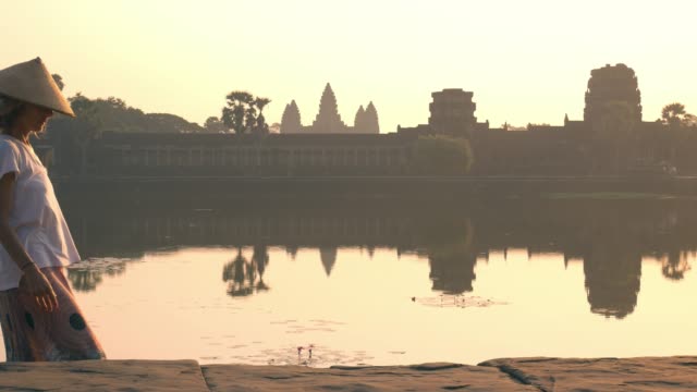 Ruinas-de-mujer-visitando-Angkor-Wat-al-amanecer,-famoso-destino-Camboya.-Reflexión-sobre-agua-y-sunburst,-luz-suave,-tonos.