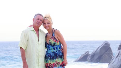 An-older-couple-smiling-on-the-beach-with-waves-and-rocks-behind-them