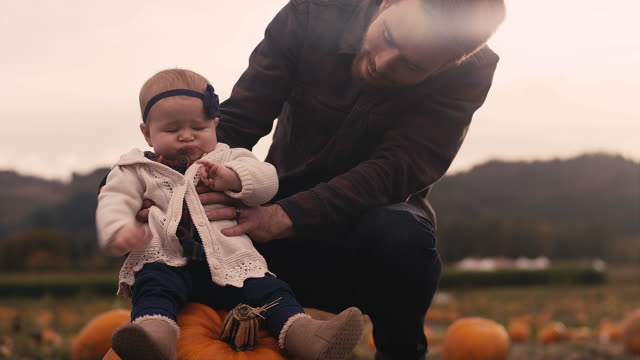 A-baby-sitting-on-a-pumpkin-at-a-pumpkin-patch,-with-her-dad-holding-her-up