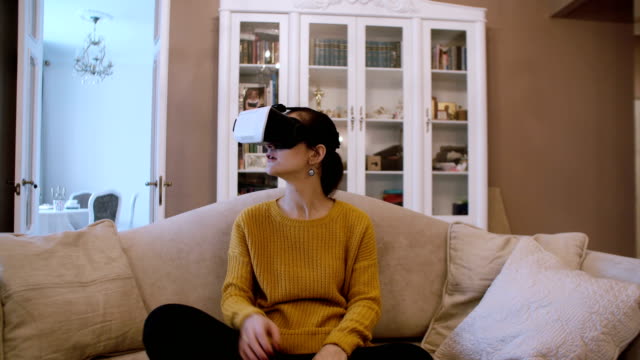 Hermosa-mujer-joven-sentada-en-el-sofá-en-la-sala-de-estar-y-utilizar-auricular-VR.-4K