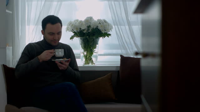Man-drinking-a-coffee-on-sofa-and-looking-through-window-at-home