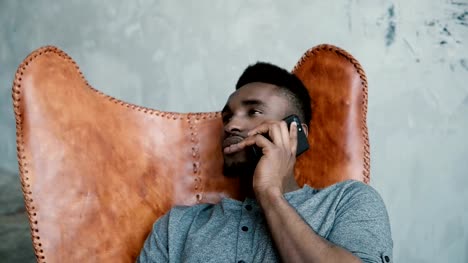 Retrato-de-africano-joven-sentado-en-la-silla,-con-Smartphone.-Un-apuesto-hombre-sonriendo-y-hablando-por-teléfono