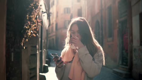 Portrait-of-young-happy-woman-walking-in-city-and-using-smartphone.-Girl-browse-the-Internet-and-drinking-coffee