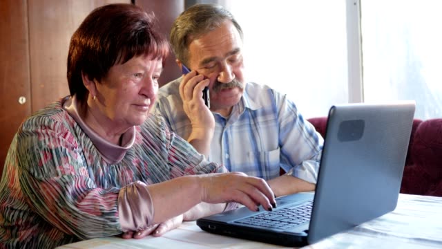An-elderly-couple-is-sitting-at-home-at-the-laptop.-Woman-reading-news,-man-talking-on-mobile-phone
