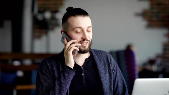 An-adult-man-with-a-beard-on-his-face,-put-his-hair-in-the-tail,-talking-on-his-mobile-phone-with-a-business-partner-in-a-restaurant-until-he-was-brought-dinner