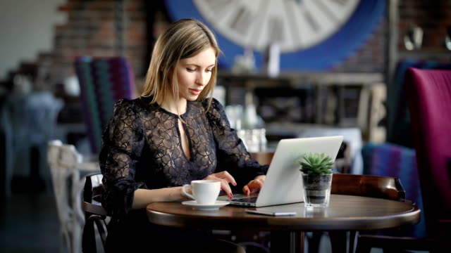 Hermosa-mujer-en-vestido-sexy-negro-sentado-en-la-mesa-en-el-restaurante-con-portátil-moderno.-Blogger-de-belleza-famoso-trabajando-en-oficina-móvil-en-la-cafetería-charlando-con-seguidores-en-dispositivos-digitales-de