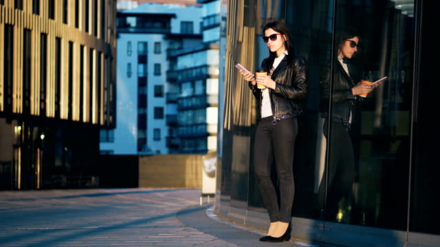 Junge-attraktive-Geschäftsfrau-mit-Tasse-Kaffee-ist-in-der-Stadt-mit-Bürogebäuden-Smartphone-verwenden.