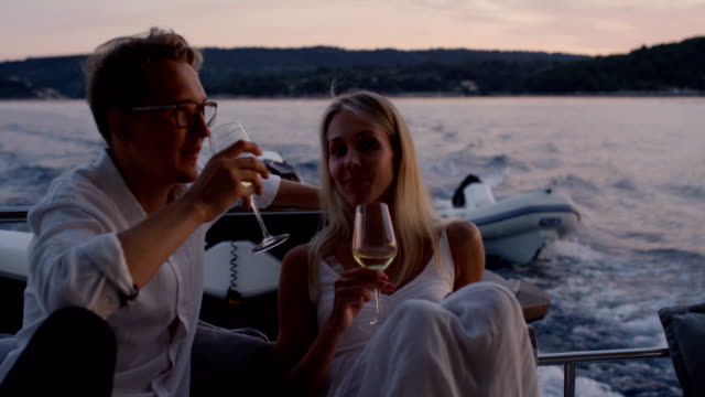 Young-Couple-Talk,-Drink-Champagne-in-the-Stern-of-the-Moving-Yacht.-They-Have-Great-Romantic-Evening.-In-the-Background-Island-with-Small-Village.