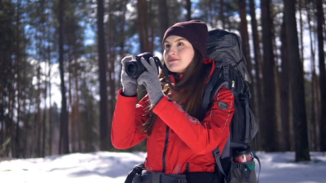 Smiling-happy-young-woman-hiking-in-winter-forest-taking-pictures-using-photocamera.