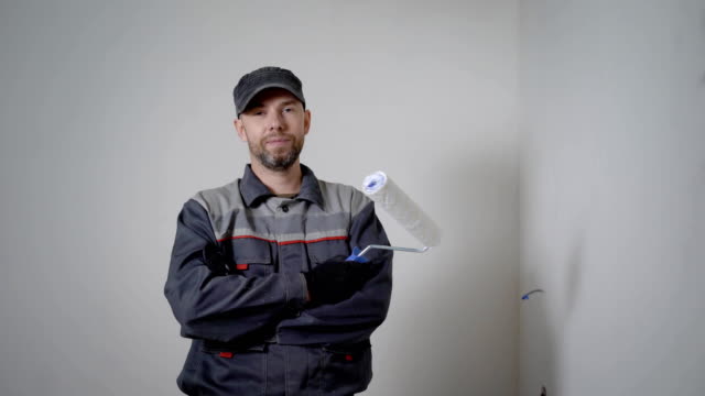 fröhliche-Mechaniker-steht-in-Wohnung,-hält-Farbroller,-Kreuzung-Hände-im-Hintergrund-weiße-leere-Wände
