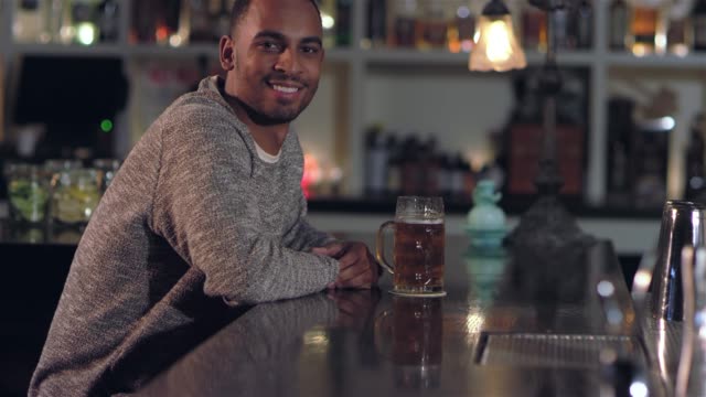 Portrait-shot-of-a-young-attractive-African-American-man-smiling,-and-taking-a-sip-of-his-beer-at-a-bar